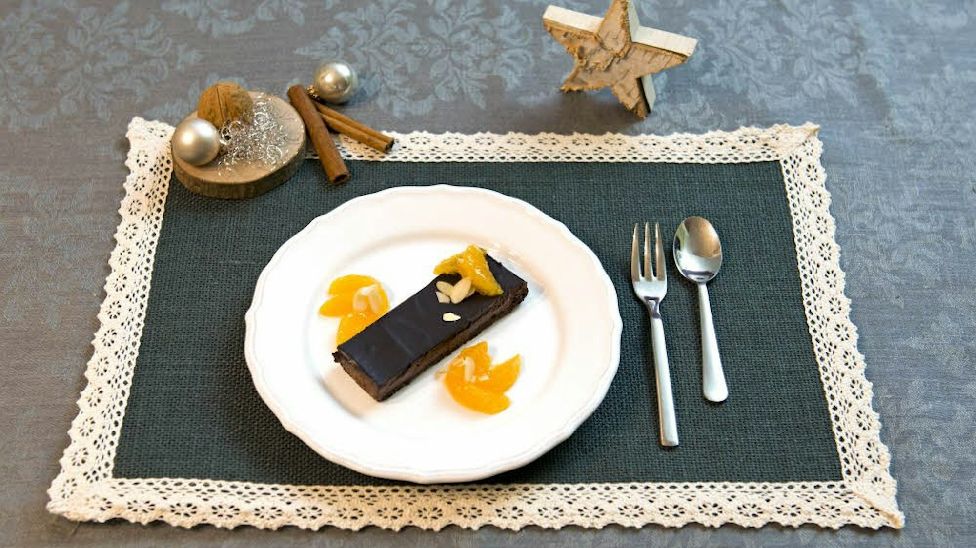 Ein Stück Schokotorte liegt, garniert mit einigen Mandarinenstücken, auf einem Teller, angerichtet auf einem Platzdeckchen mit weihnachtlicher Deko.