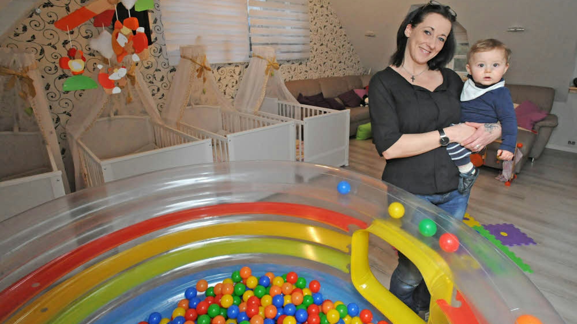 Tagesmutter mit Kind auf dem Arm steht neben einem Bällchen-Bad im Spielzimmer.