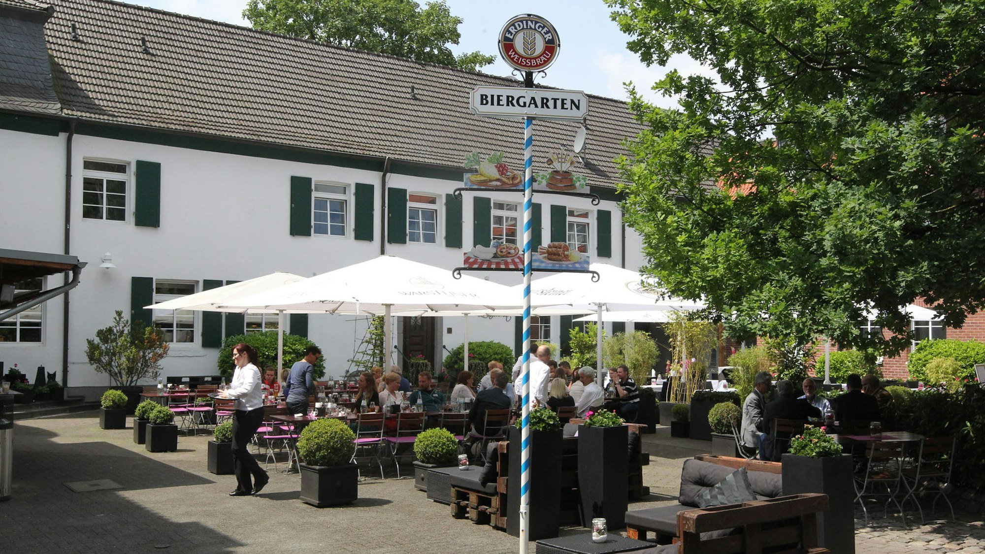 Vor einem weißen Haus stehen Tische und Stühle unter Sonnenschirmen. Ein Biergarten-Schild steht auf dem Platz.