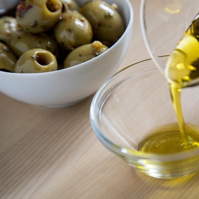 Aus einer Glaskaraffe wird Olivenöl in eine Glasschale gegossen, im Hintergrund eine Porzellanschale mit grünen Oliven.