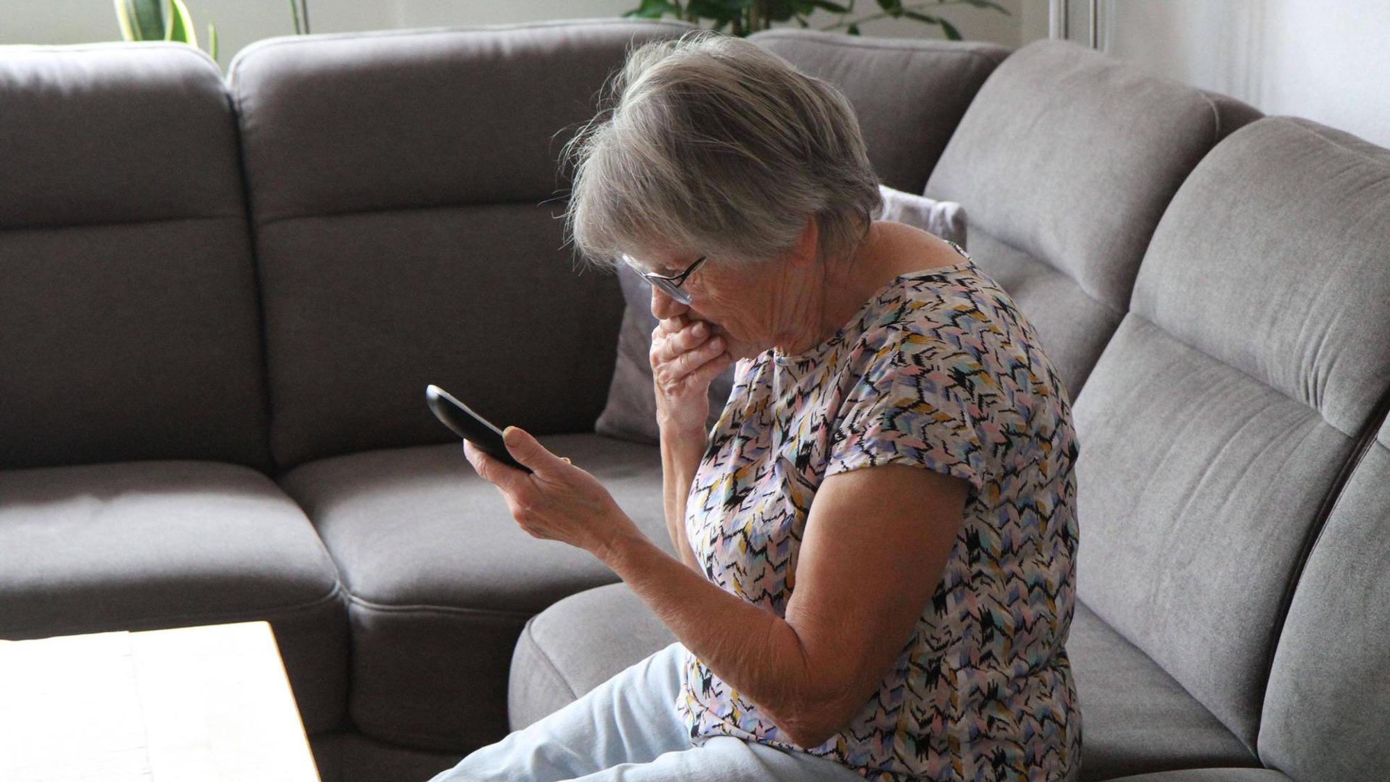 Das Foto zeigt einen schockiert wirkende Frau auf einem Sofa, die ein Telefon in der Hand hält.
