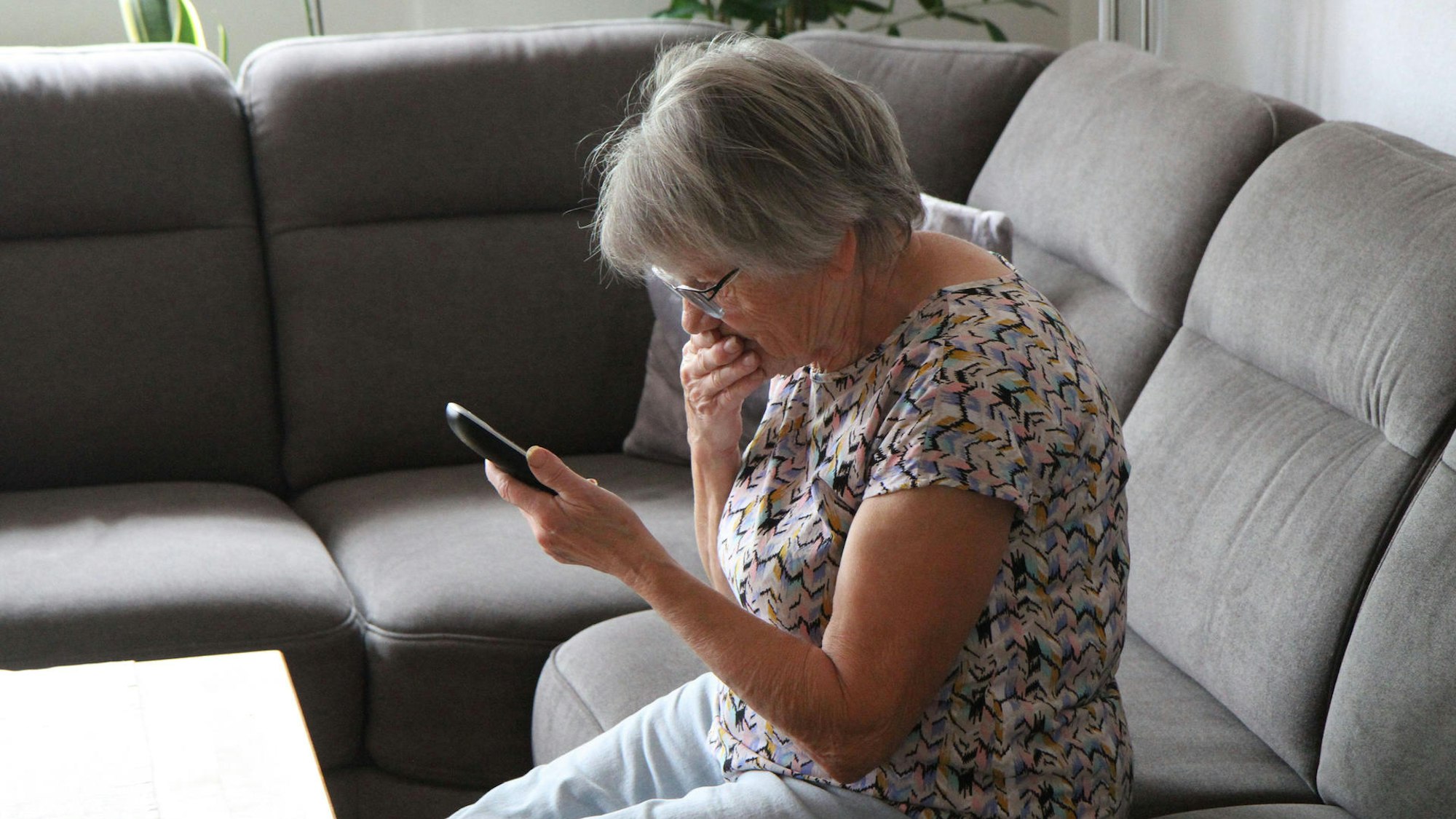 Das Foto zeigt einen schockiert wirkende Frau auf einem Sofa, die ein Telefon in der Hand hält.