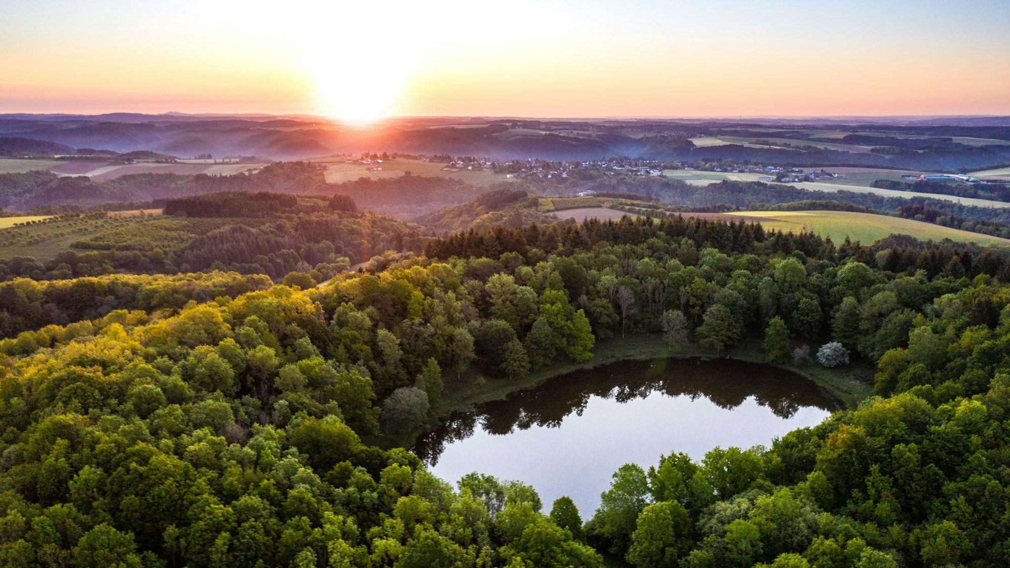 Blick aus der Luft auf den Windsborn Kratersee, rundherum wachsen Bäume, am Horizont steht die Sonne