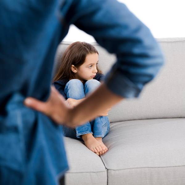 Mädchen sitzt auf der Couch und dreht sich weg, ein Erwachsenen von hinten, der die Hände in die Hüften stützt.