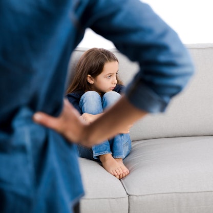 Mädchen sitzt auf der Couch und dreht sich weg, ein Erwachsenen von hinten, der die Hände in die Hüften stützt.