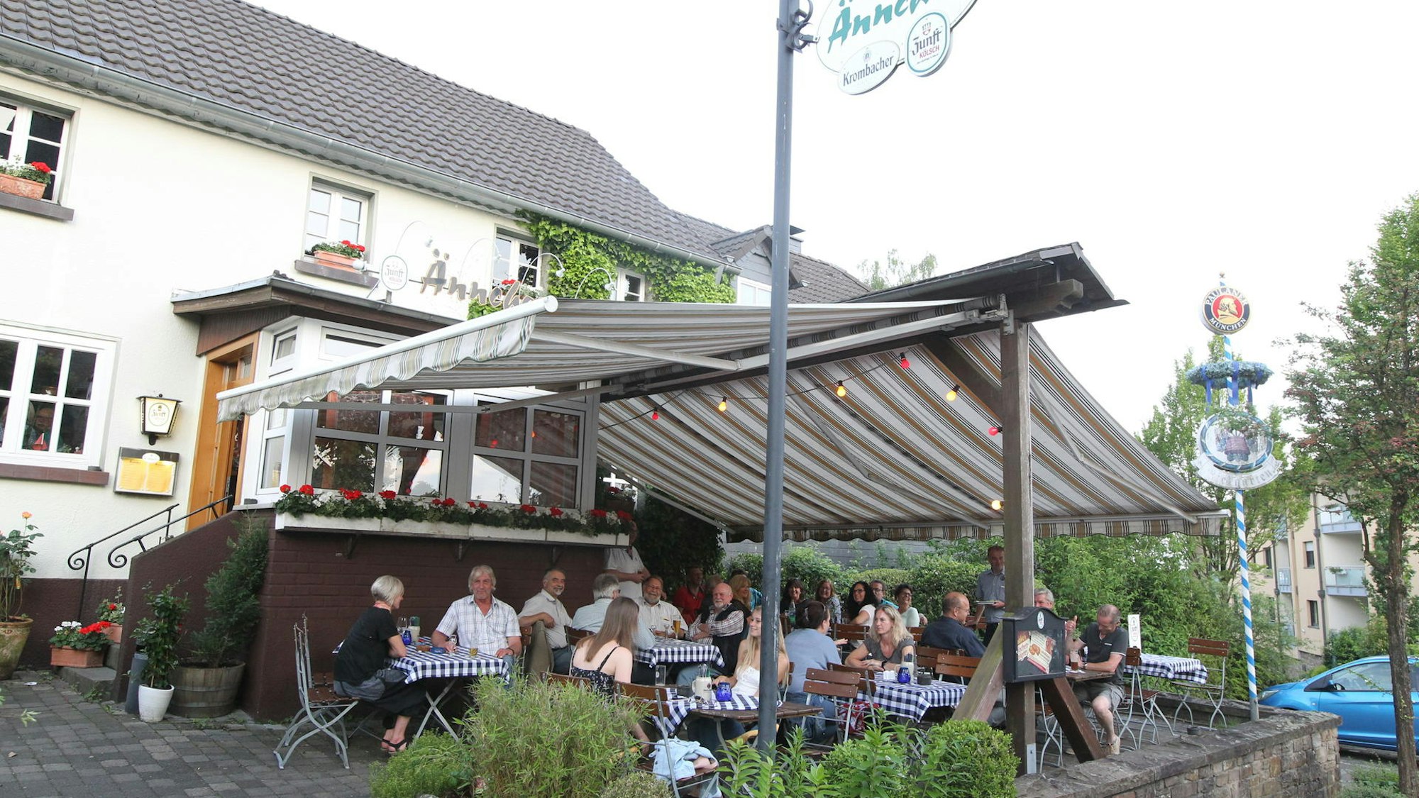 Unter einer Marquise sitzen mehrere Menschen an Tischen vor einem Gebäude, auf dem „Ännchen“ steht.