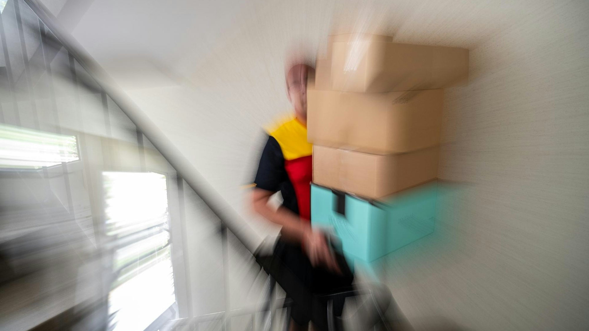 Ein Paketzusteller im Einsatz: Ein Mann mit rot-gelbem T-Shirt trägt vier große Pakete eine Treppe hinauf.