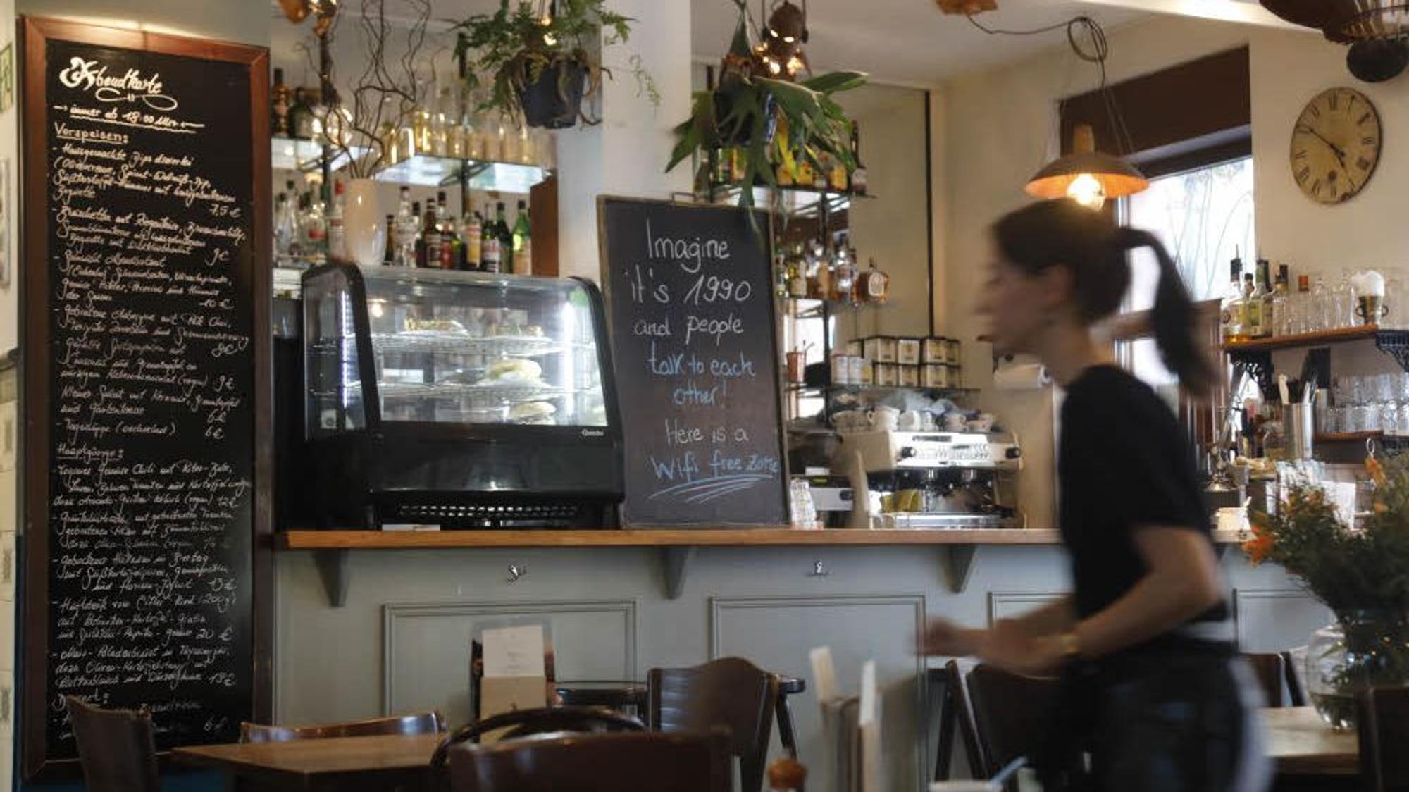 Das Café Sehnsucht in Köln von innen, mit zwei Tafeln mit dem aktuellen Angebot und einer geblurrten Thekenkraft.
