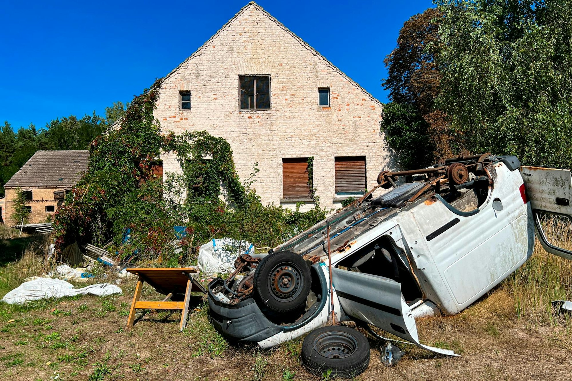 Ein altes Auto liegt auf dem Kopf vor einem Verlassenen Haus.