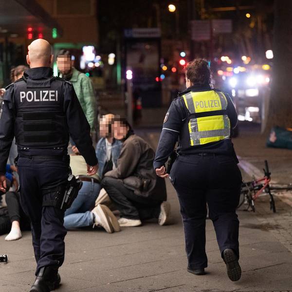 Menschen hocken nachts auf dem Hohenzollernring im Kreis um einen Verletzten, der niedergeschlagen wurde, zwei Polizisten eilen hinzu.