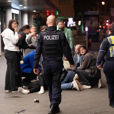 Menschen hocken nachts auf dem Hohenzollernring im Kreis um einen Verletzten, der niedergeschlagen wurde, zwei Polizisten eilen hinzu.