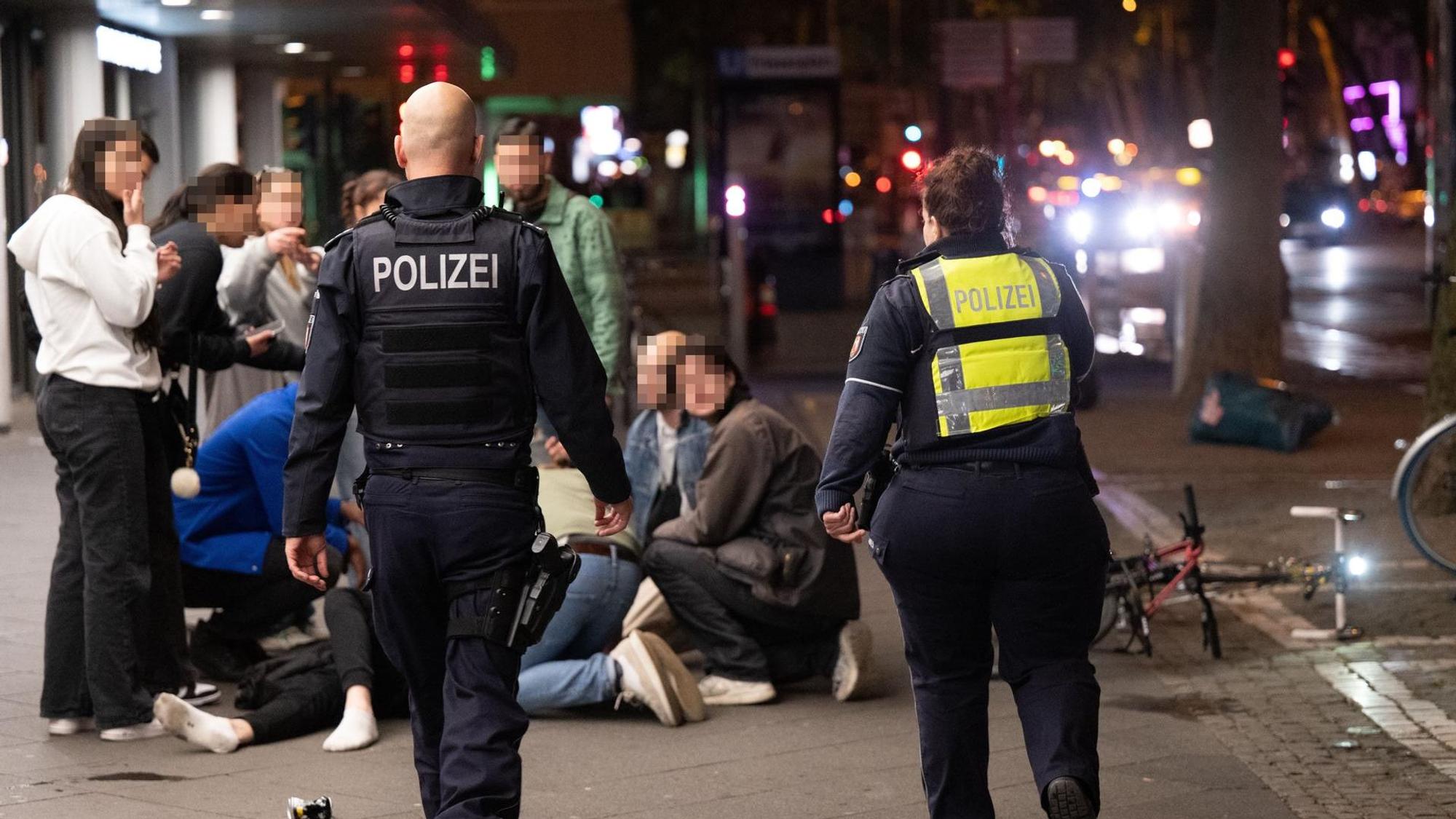Menschen hocken nachts auf dem Hohenzollernring im Kreis um einen Verletzten, der niedergeschlagen wurde, zwei Polizisten eilen hinzu.