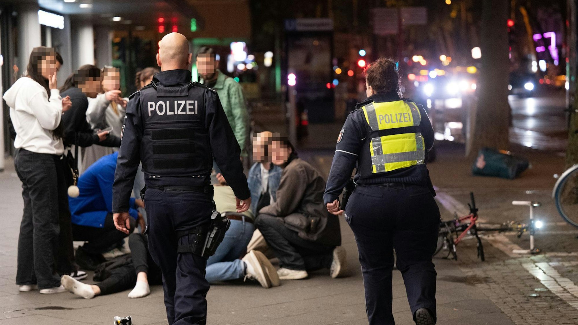 Menschen hocken nachts auf dem Hohenzollernring im Kreis um einen Verletzten, der niedergeschlagen wurde, zwei Polizisten eilen hinzu.