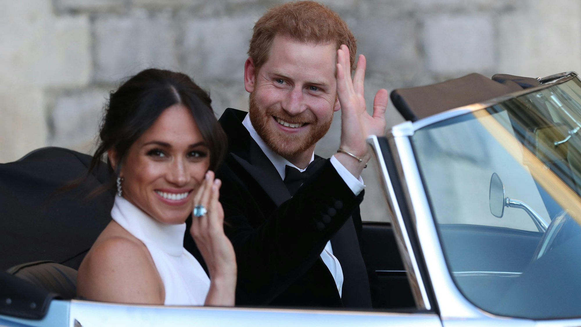 Meghan und Harry winken in einem Auto.