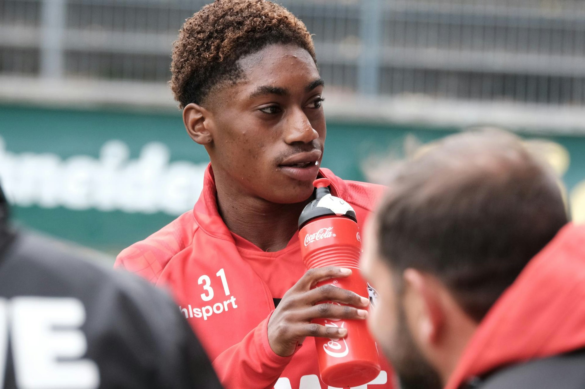 Ein Fußballspieler steht mit seiner Trinkflasche in der Hand am Rande des Platzes.