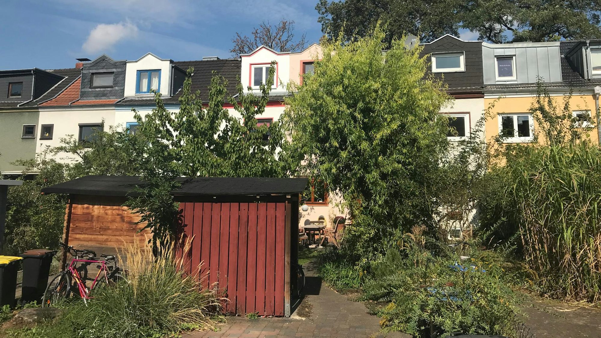 Ein roter Schuppen in einem Garten mit kleinen Bäumen, dahinter eine Zeile kleiner Reihenhäuser.