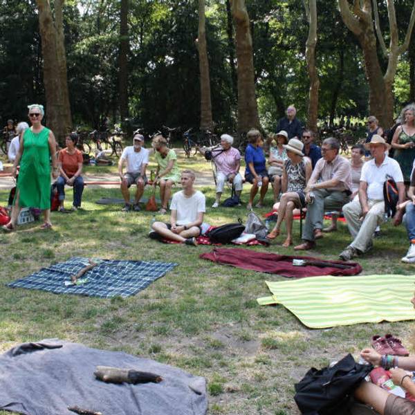Rund um eine Reihe ausgebreiterte Picknick-Decken sitzen Menschen im Kreis, im Hintergrund eine Baumreihe. Eine Frau redet in ein Mikrofon.