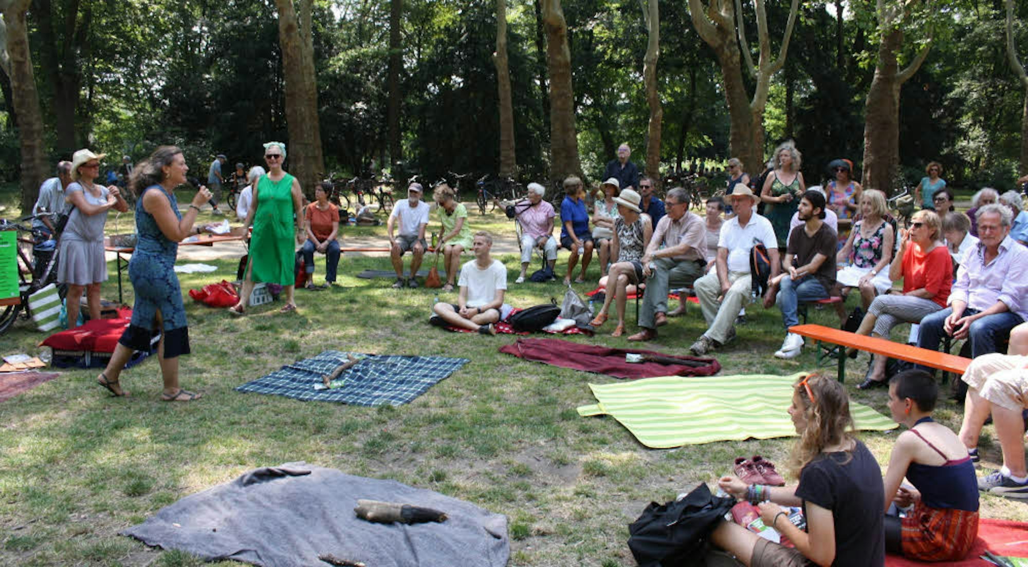 Rund um eine Reihe ausgebreiterte Picknick-Decken sitzen Menschen im Kreis, im Hintergrund eine Baumreihe. Eine Frau redet in ein Mikrofon.