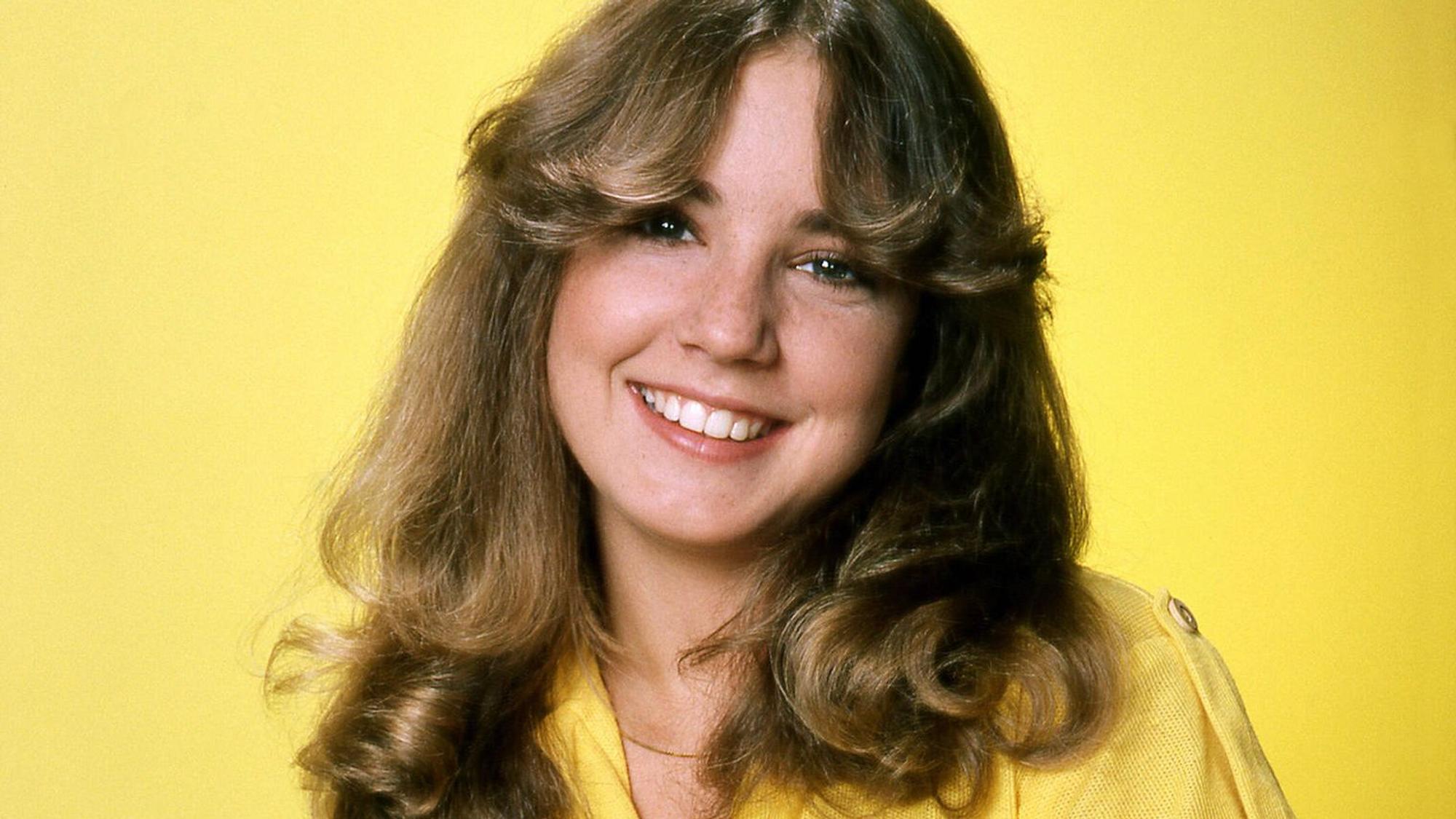 Dana Plato mit 70er-Jahre-Föhnwelle und gelben Outfit vor einem gelben Hintergrund. Dana Plato lacht freundlich in die Kamera.