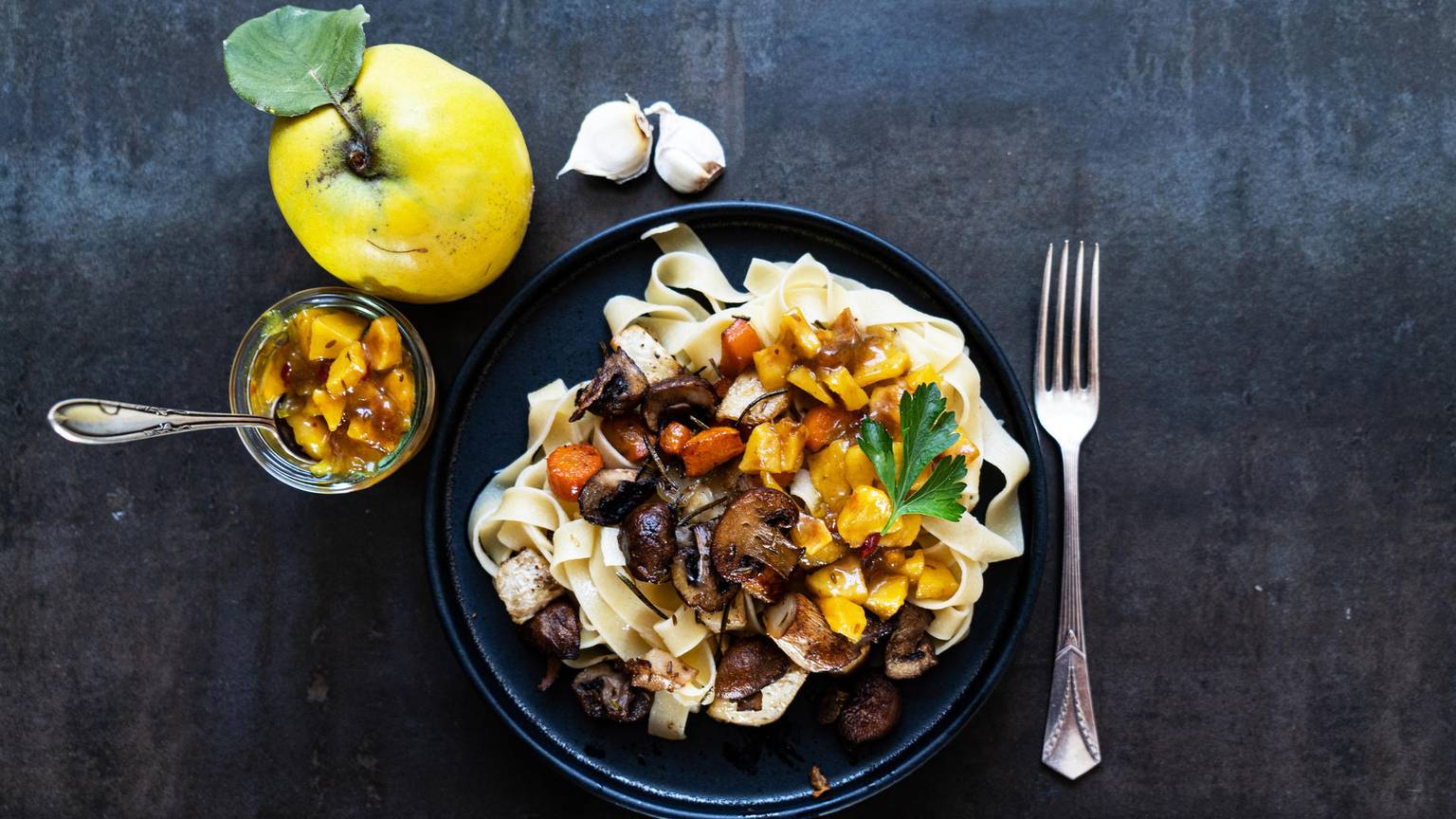 auf einem Teller liegen Fettucine, darauf Gemüse und Quitten-Stücke, links daneben liegt eine Quitte und ein Glas mit Quitten-Chutney