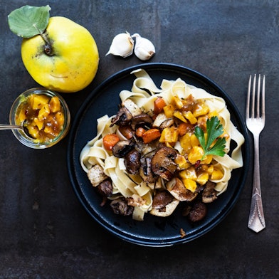 auf einem Teller liegen Fettucine, darauf Gemüse und Quitten-Stücke, links daneben liegt eine Quitte und ein Glas mit Quitten-Chutney
