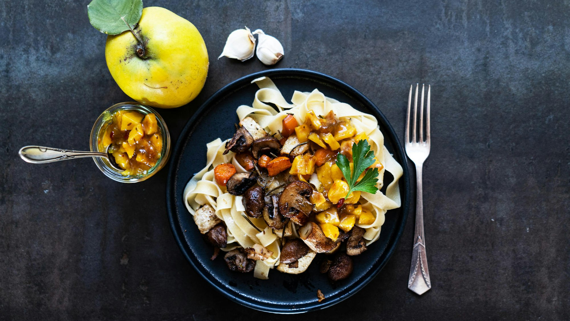 auf einem Teller liegen Fettucine, darauf Gemüse und Quitten-Stücke, links daneben liegt eine Quitte und ein Glas mit Quitten-Chutney