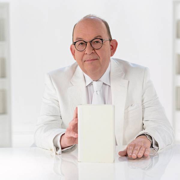 Denis Scheck sitzt in einem weißen Anzug in einem Studio an einem weißen Schreibtisch und hat ein weißes Buch in der Hand.