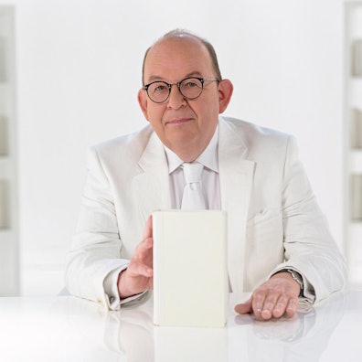 Denis Scheck sitzt in einem weißen Anzug in einem Studio an einem weißen Schreibtisch und hat ein weißes Buch in der Hand.