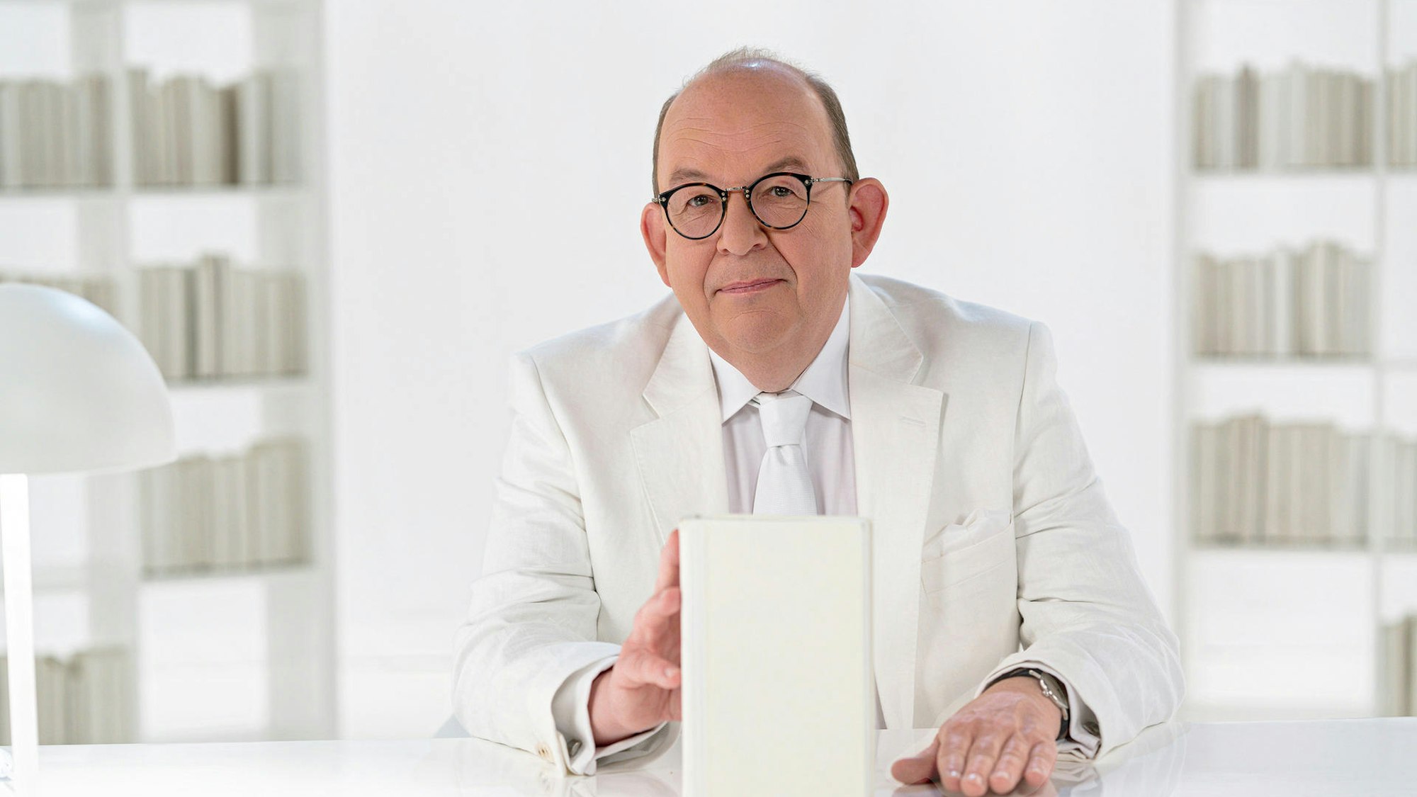 Denis Scheck sitzt in einem weißen Anzug in einem Studio an einem weißen Schreibtisch und hat ein weißes Buch in der Hand.