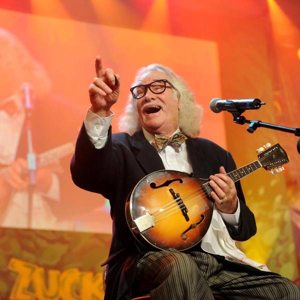 Hans Süper sitzt lachend mit Mandoline auf einer Bühne und zeigt ins Publikum.
