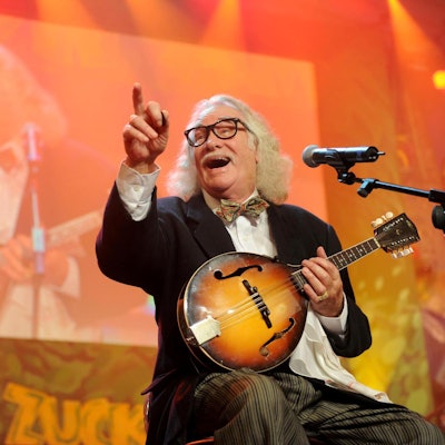 Hans Süper sitzt lachend mit Mandoline auf einer Bühne und zeigt ins Publikum.