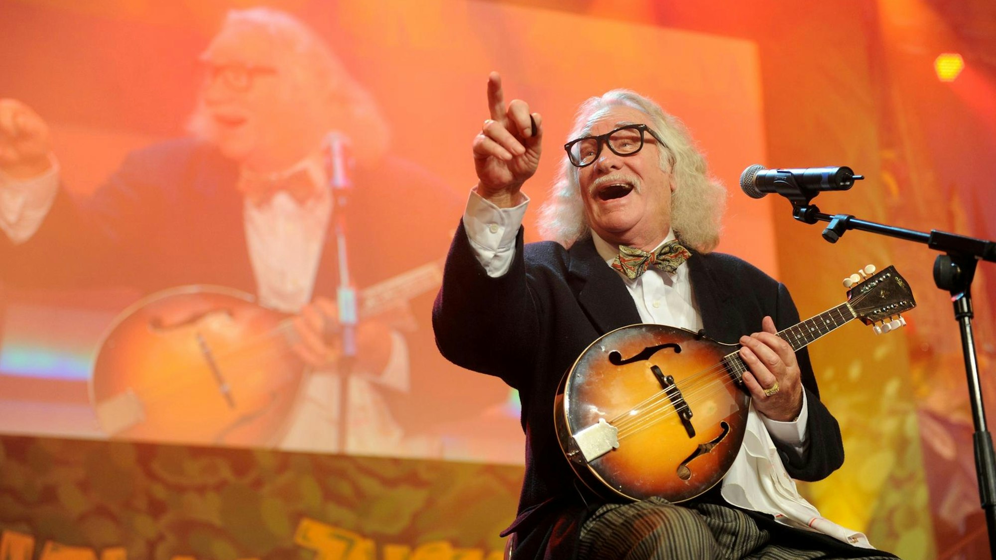 Hans Süper sitzt lachend mit Mandoline auf einer Bühne und zeigt ins Publikum.