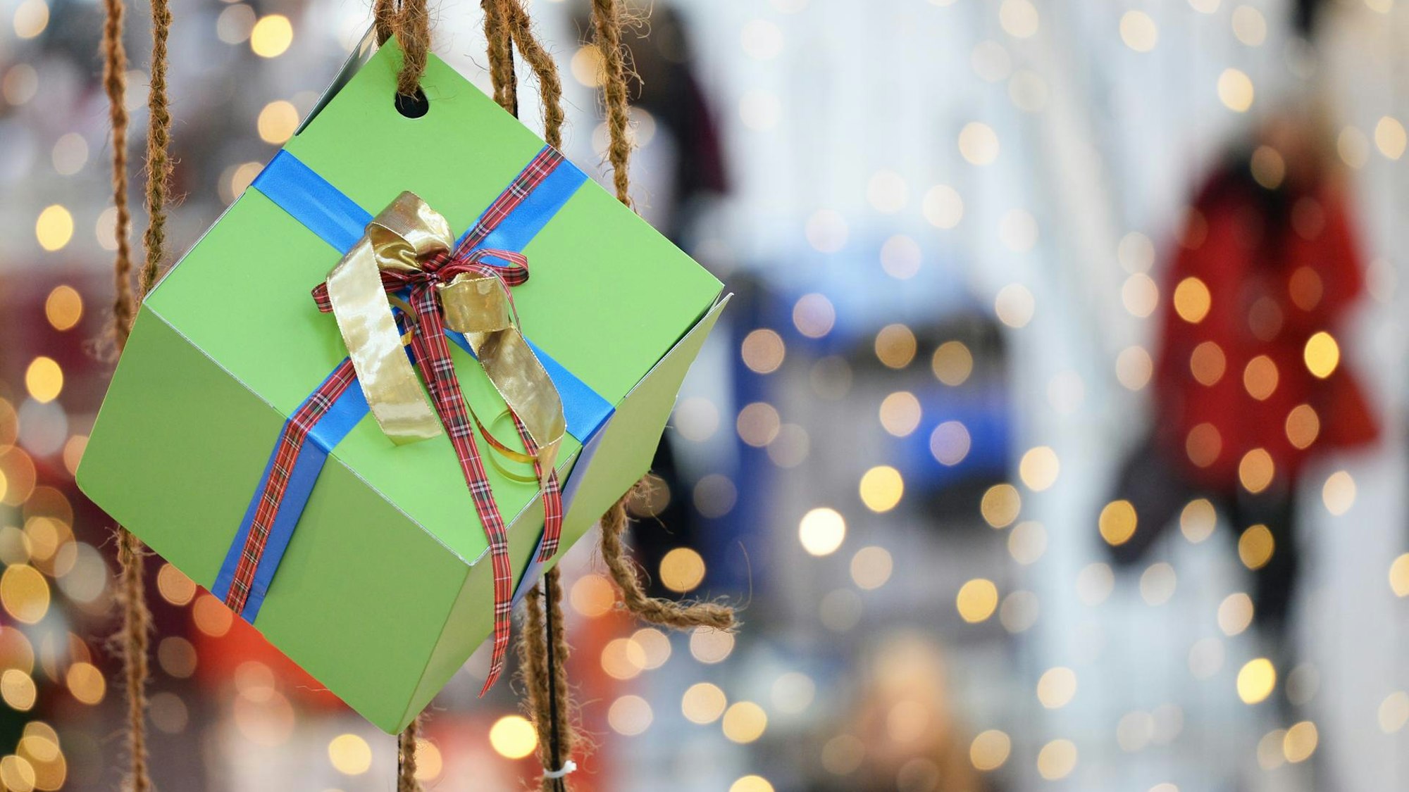 Ein kleines grünes Päckchen mit roter, blauer und goldener Schleife hängt an einem Band vor verschwommenem Hintergrund
