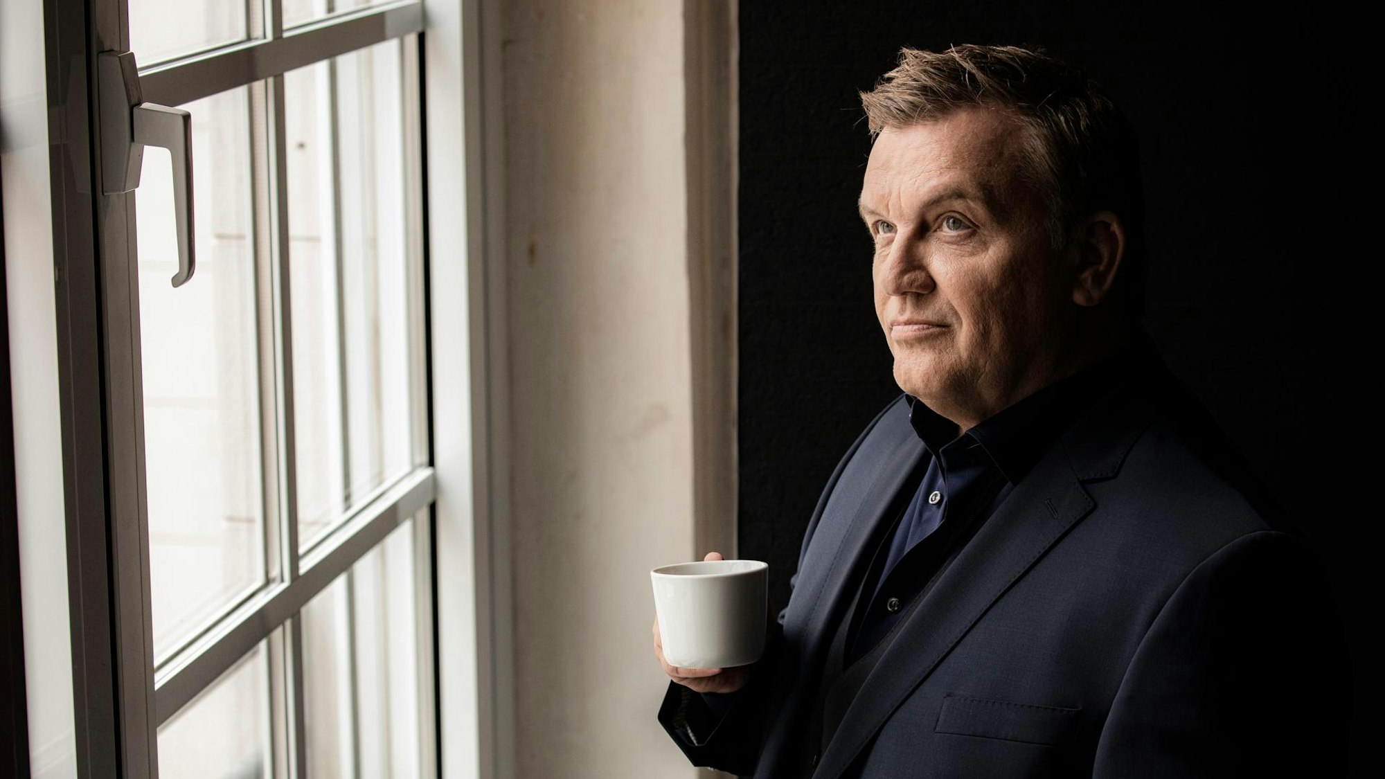 Hape Kerkeling steht mit einer Kaffeetasse an einem Fenster und schaut nachdenklich.