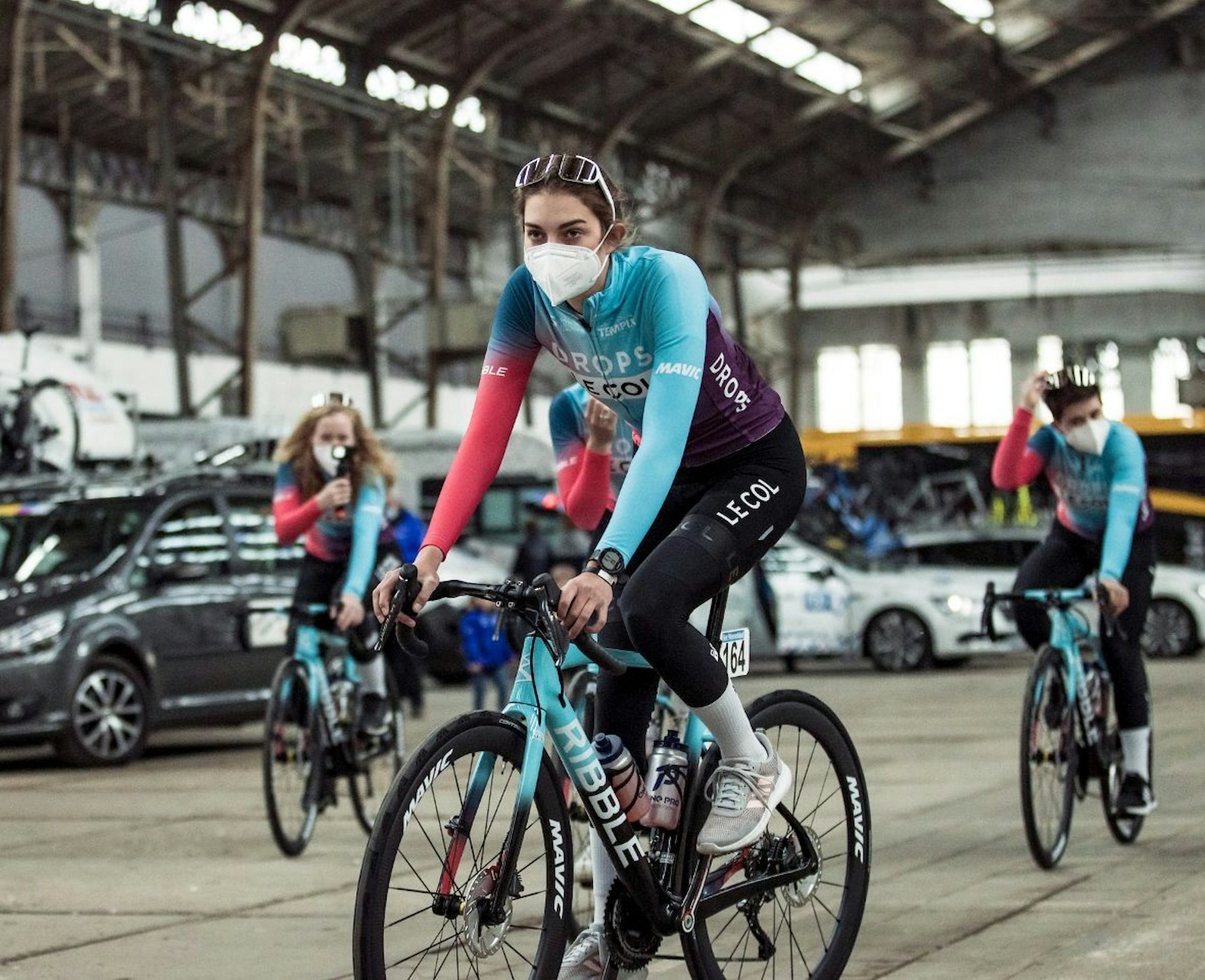 Finja Smelka fährt mit ihrem Fahrrad durch eine Halle. Im Hintergrund fahren noch drei weitere Personen auf einem Fahrrad hinter ihr her. Eine Frau hält eine kleine Kamera in der Hand.
