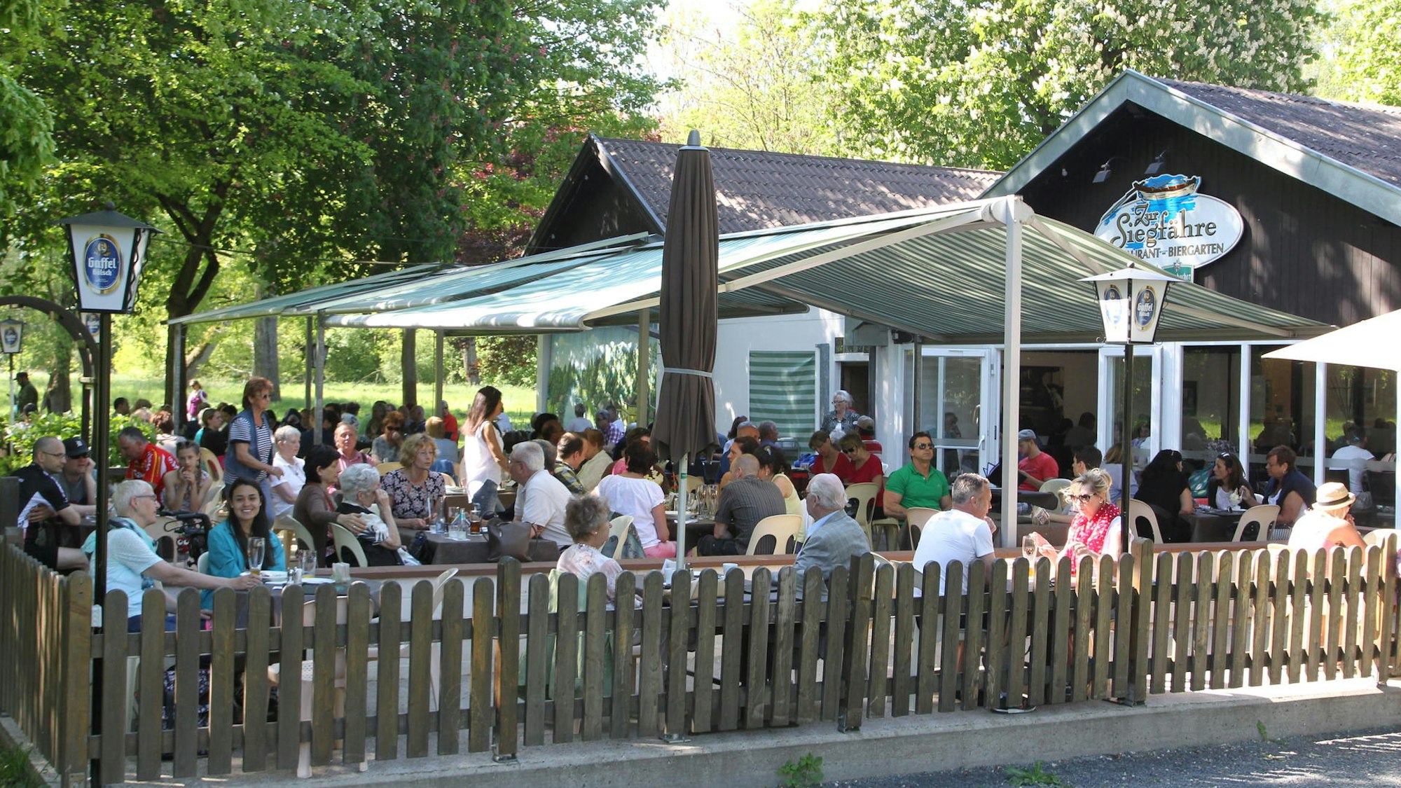 Vor einem Gebäude stehen Bänke und Tischen im Schatten. Viele Personen sitzen dort.
