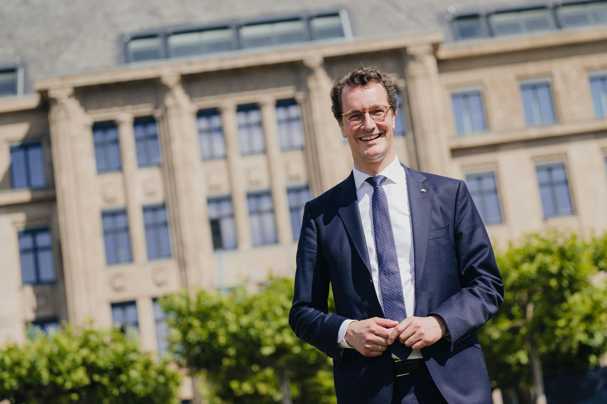 Hendrik Wüst (CDU) steht vor der historische Staatskanzlei in Düsseldorf, er ist im Halbporträt zu sehen vor der Fassade und einer grünen Baumreihe im Sonnenschein. Er lacht in die Kamera.
