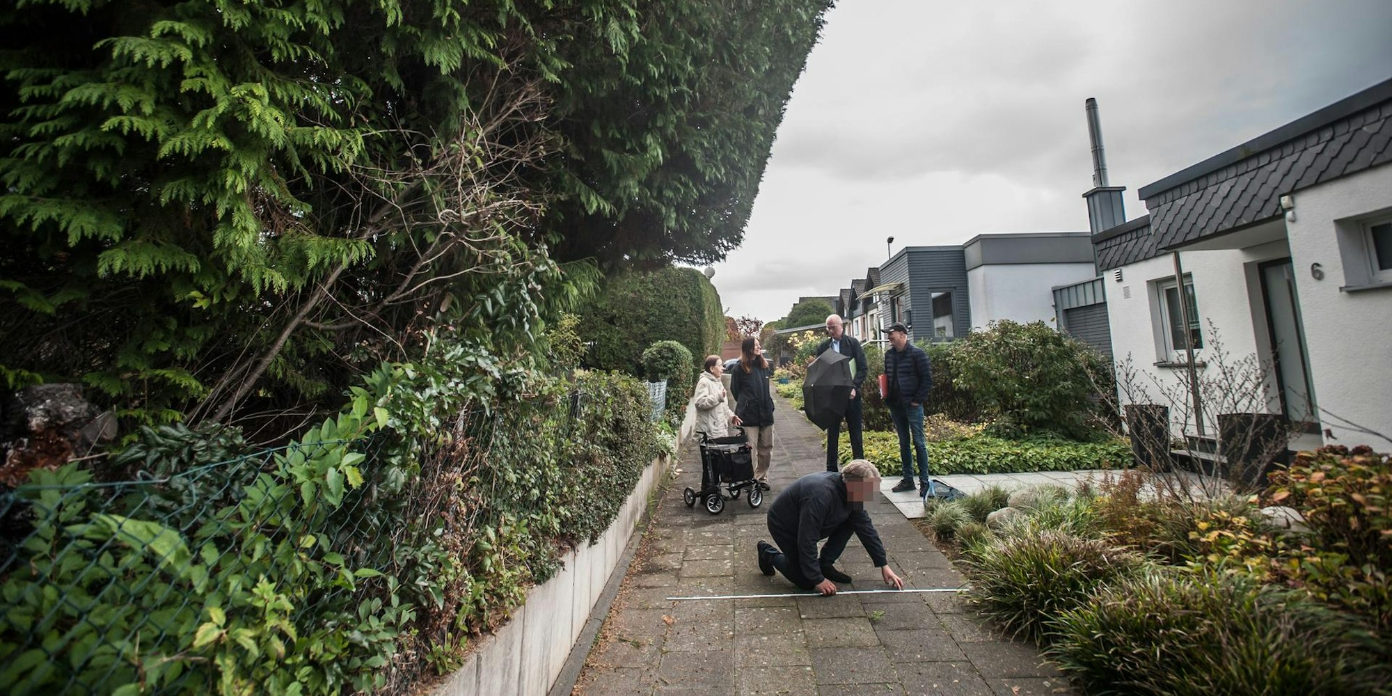 Mehrere Personen stehen auf einem Steinweg. Rechts befinden sich Wohnhäuser, links Pflanzen und Bäume. Auf dem Boden kniet ein Mann und misst die Breite des Gehwegs.