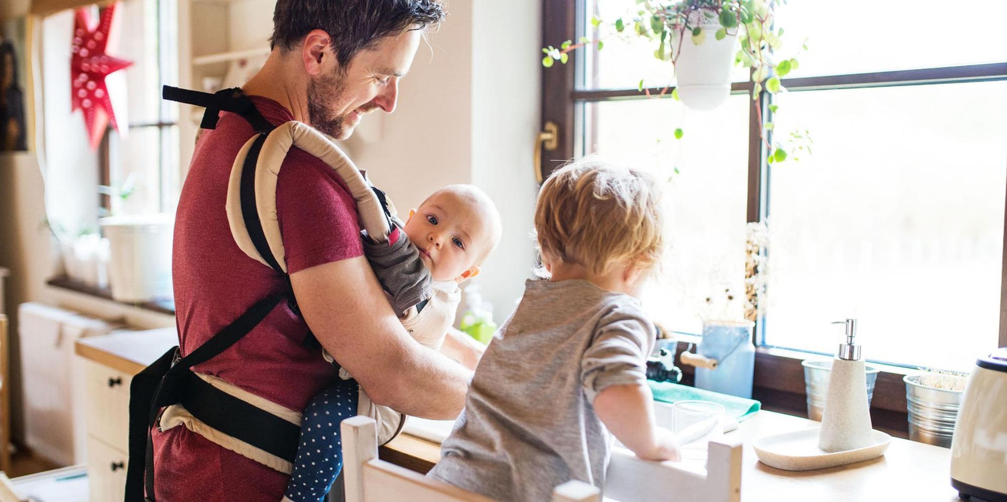 Ein Mann hat ein Baby in der Trage und spült ab.