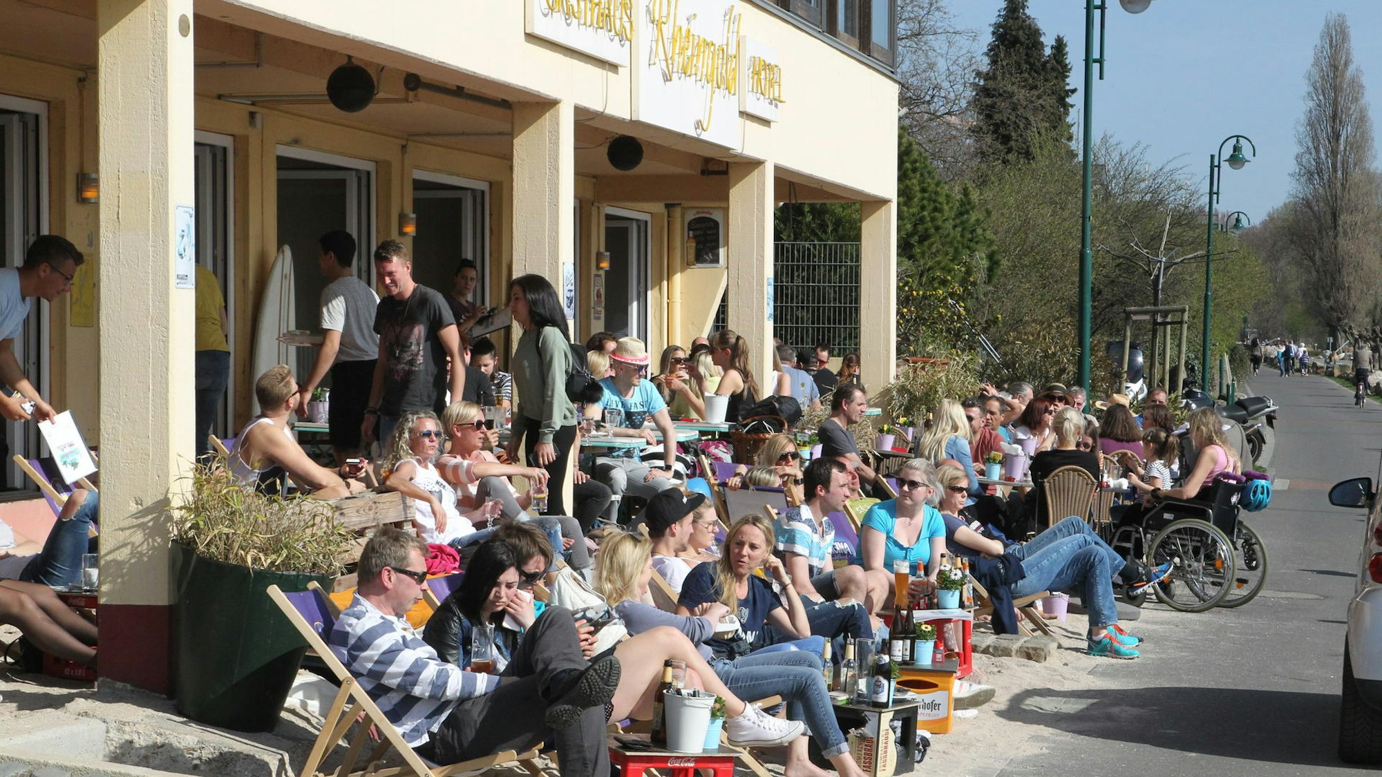 Viele Personen sitzen vor einem Gebäude in Liegestühlen oder auf Stühlen. Am Boden ist Sand verteilt.