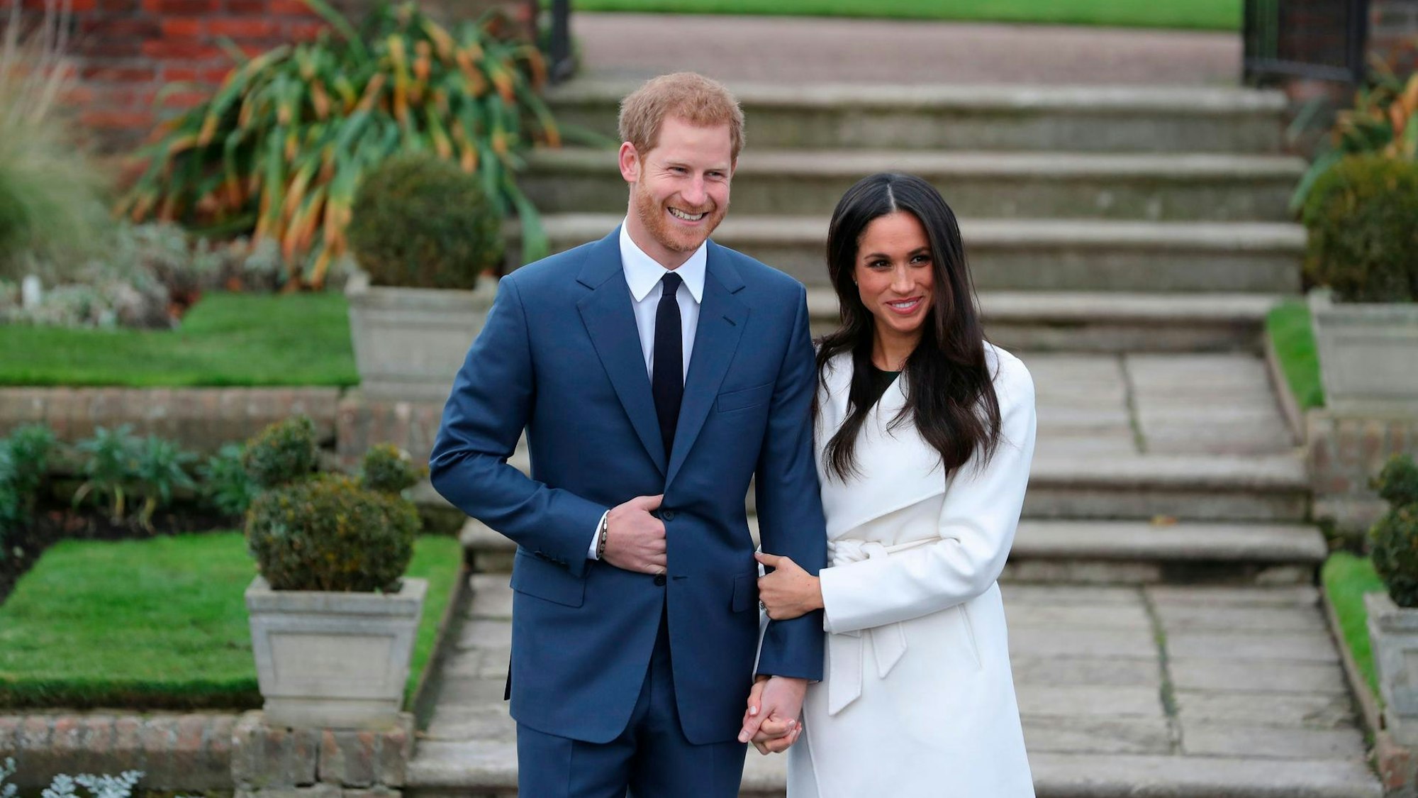 Harry und Meghan bei einem Fototermin. Sie halten Hand und lächeln freundlich.