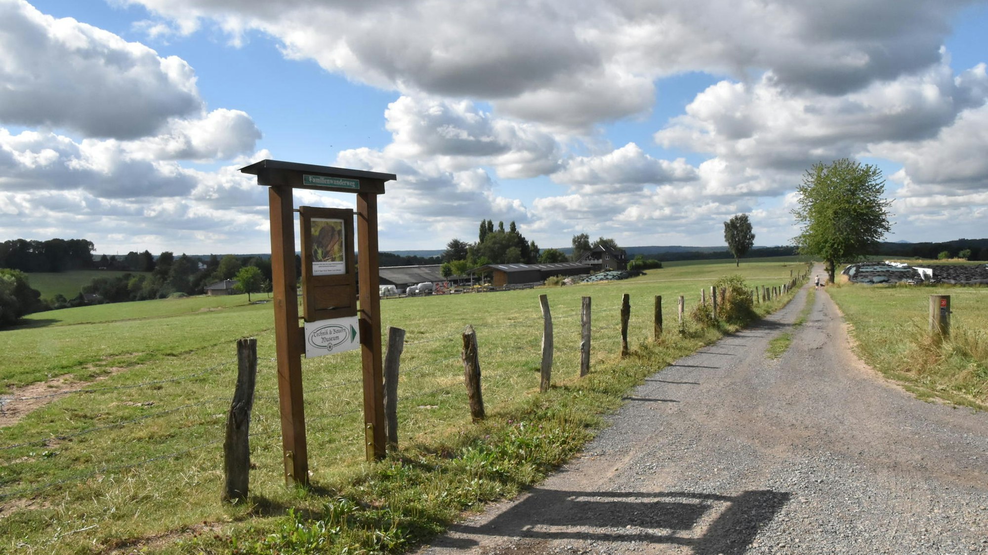Ein Schild steht am Rand eines Wanderwegs. Er verläuft zwischen Wiesen.