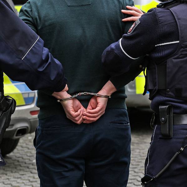 Zwei Polizisten führen einen Mann ab, dessen Hände auf dem Rücken gefesselt sind.