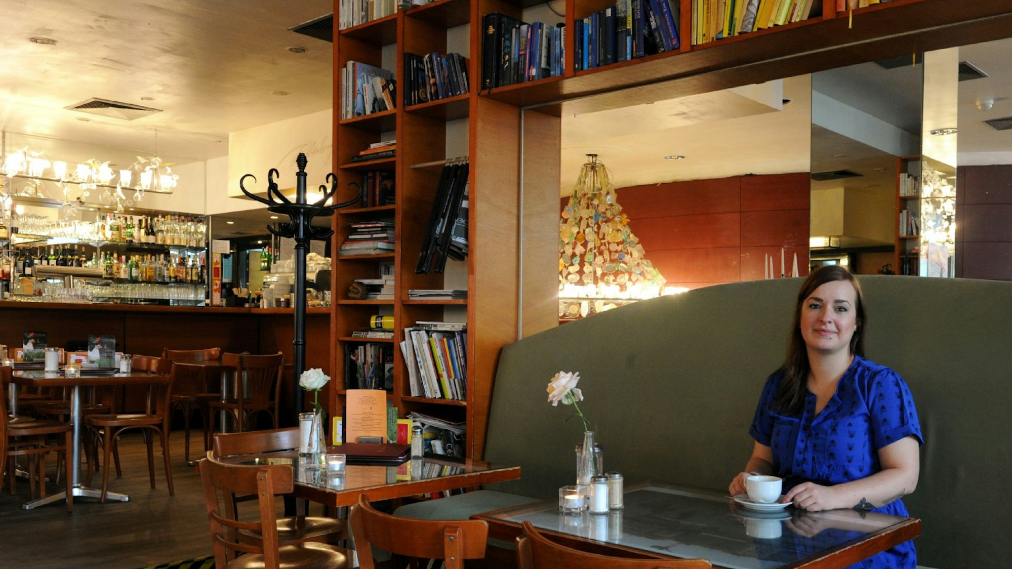 Julia Floß sitzt in einem blauen Kleid auf einem Sofa, vor ihr steht eine Tasse Kaffee, im Hintergrund ist ein Bücherregal zu sehen