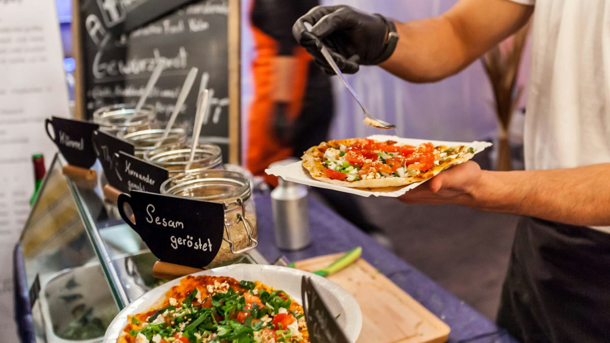 Ein Mann bereitet eine Pizza zu. Vor ihm stehen diverse Zutaten. Er streut gerade Gewürze mit einem Löffel auf die Pizza.