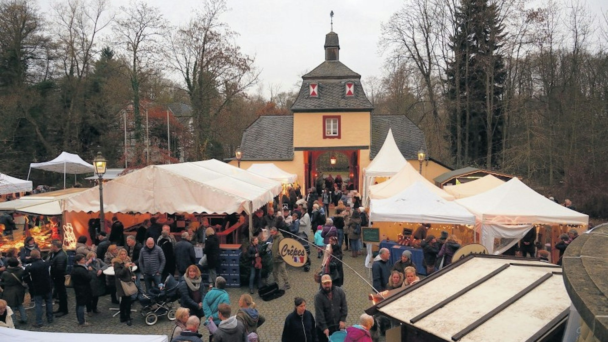 An den Buden stehen viele Menschen. Im Hintergrund liegt das Tor von Schloss Eulenbroich.