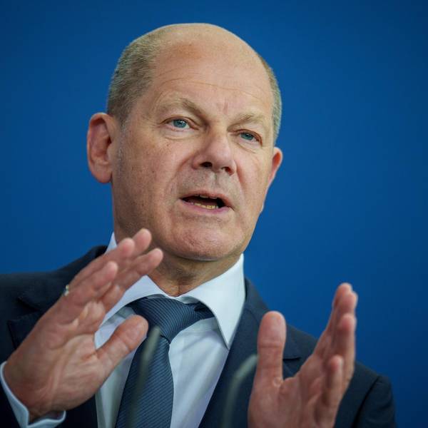 Bundeskanzler Olaf Scholz steht vor einer blauen Wand, spricht und gestikuliert mit seinen Händen.
