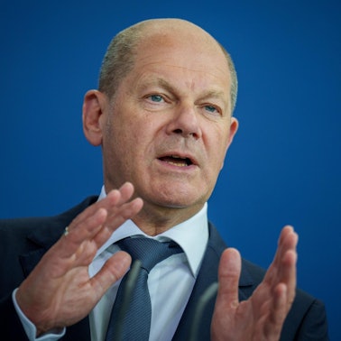 Bundeskanzler Olaf Scholz steht vor einer blauen Wand, spricht und gestikuliert mit seinen Händen.