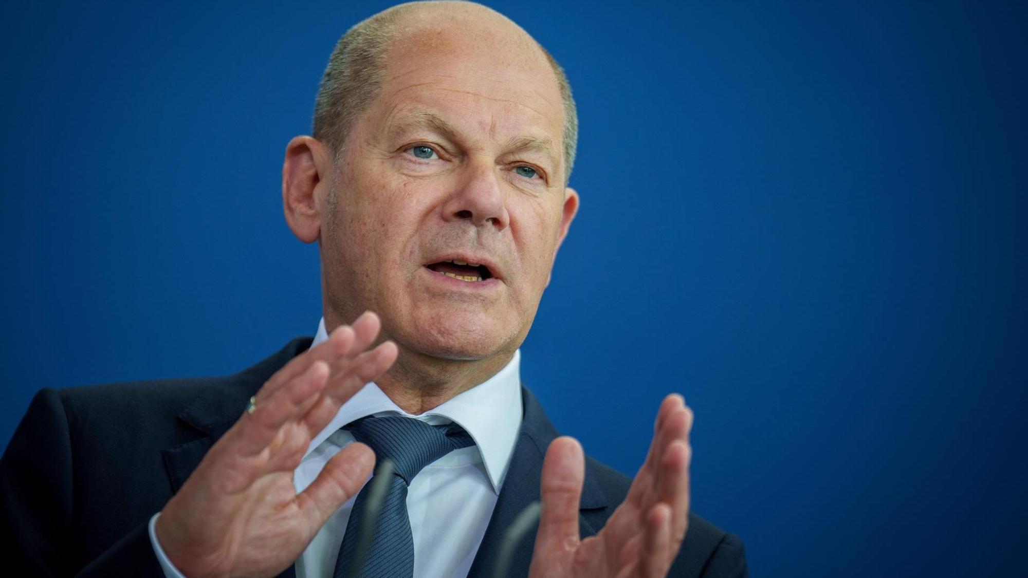 Bundeskanzler Olaf Scholz steht vor einer blauen Wand, spricht und gestikuliert mit seinen Händen.