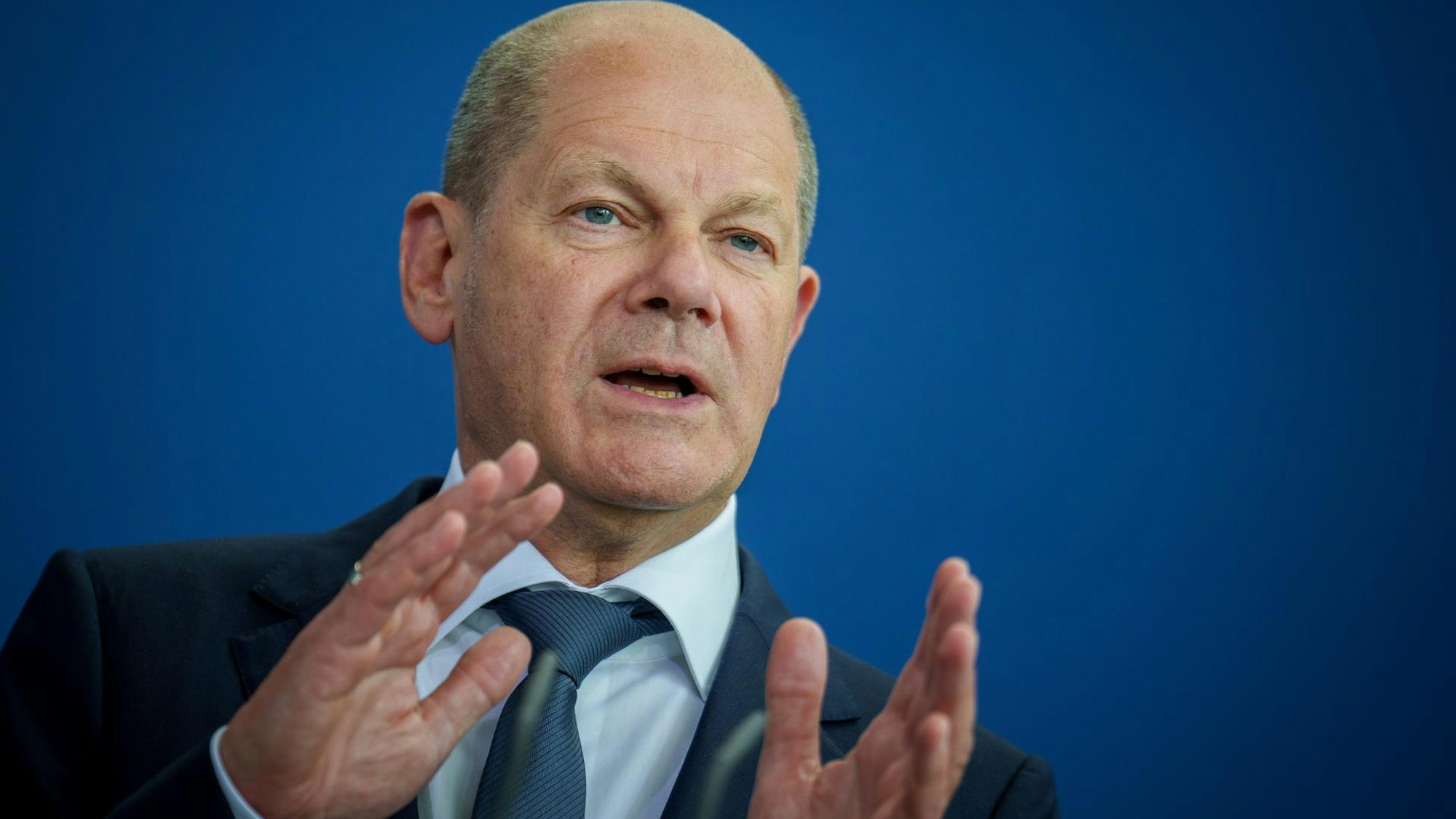Bundeskanzler Olaf Scholz steht vor einer blauen Wand, spricht und gestikuliert mit seinen Händen.